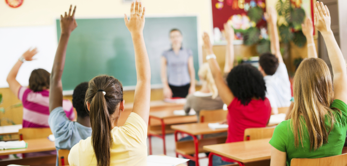 Group of teenagers sitting in classroom with raised hands.